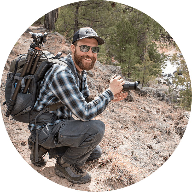 Alex Kent smiling with camera in hand crouching on a ridge overlooking a river. @alexkentphoto